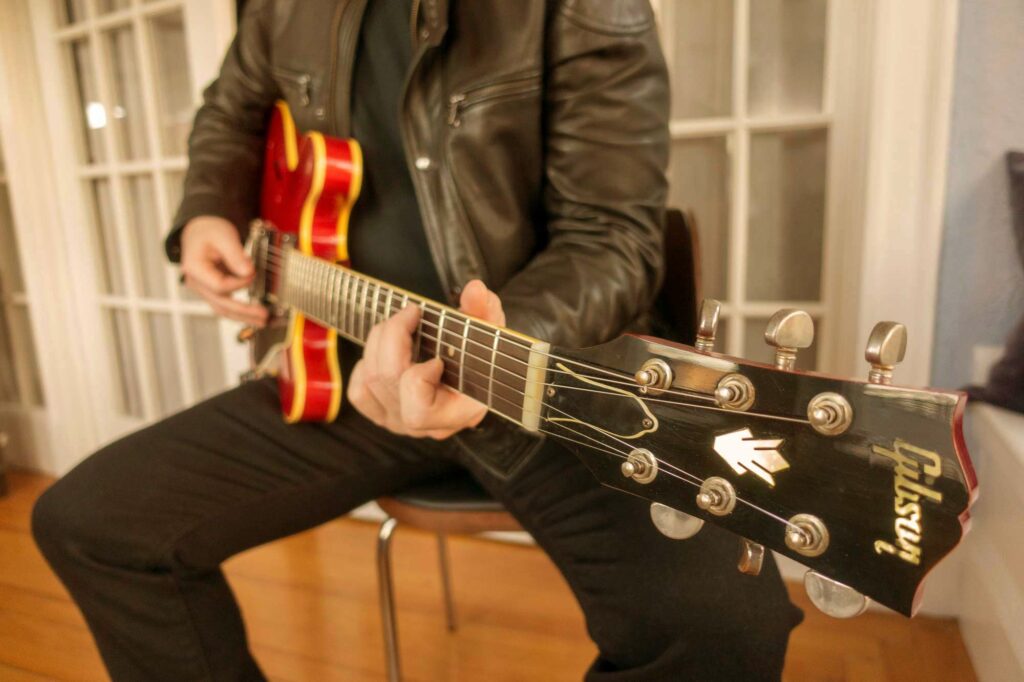 Warm, golden-lit studio portrait of a leather-jacketed guitarist playing a cherry-red Gibson semi-hollow electric.