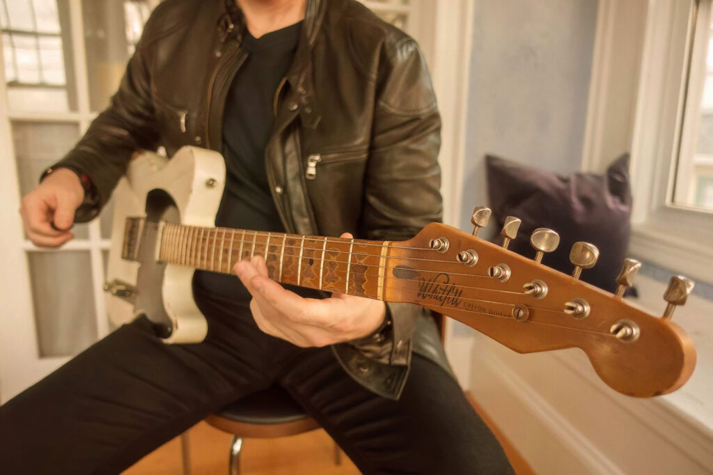 Close studio shot of a player with a road-worn cream-bodied custom electric and an aged maple neck in warm window light.