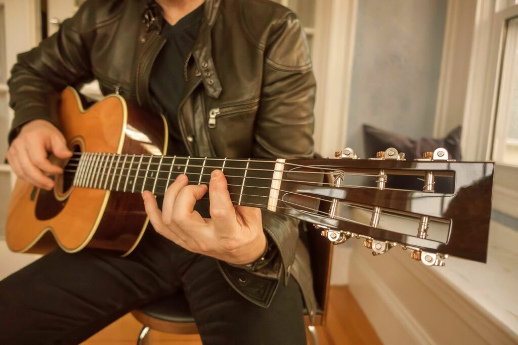 Guitarist fingerpicking a natural-top cutaway acoustic in a sunlit window seat.