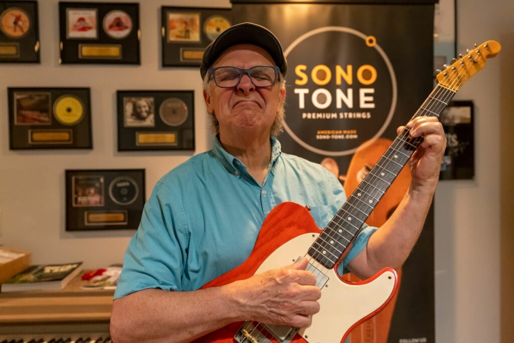 Session guitarist in a cap holding a red Telecaster before the SonoTone backdrop and a wall of gold and platinum records.