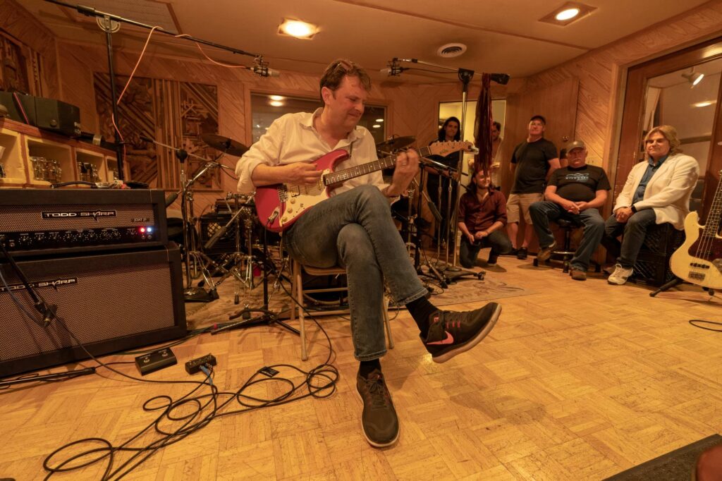 Guitarist seated playing a red Telecaster during a SonoTone recording session in a wood-paneled studio with onlookers.