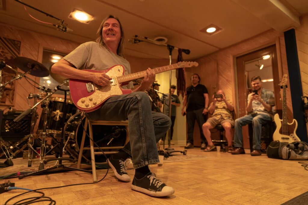 Smiling long-haired guitarist playing a pink Telecaster at a SonoTone studio session as the audience films on phones.