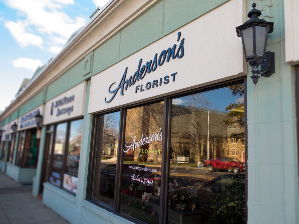 Navy script sign band above the shop windows, flanked by a lantern sconce, with window lettering and the neighborhood reflected below.