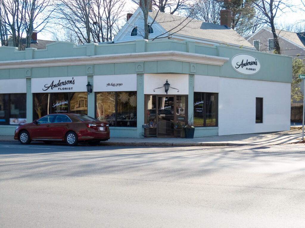 Mint-green corner storefront with the sign band and oval blade sign carrying the script mark, seen from across the street.