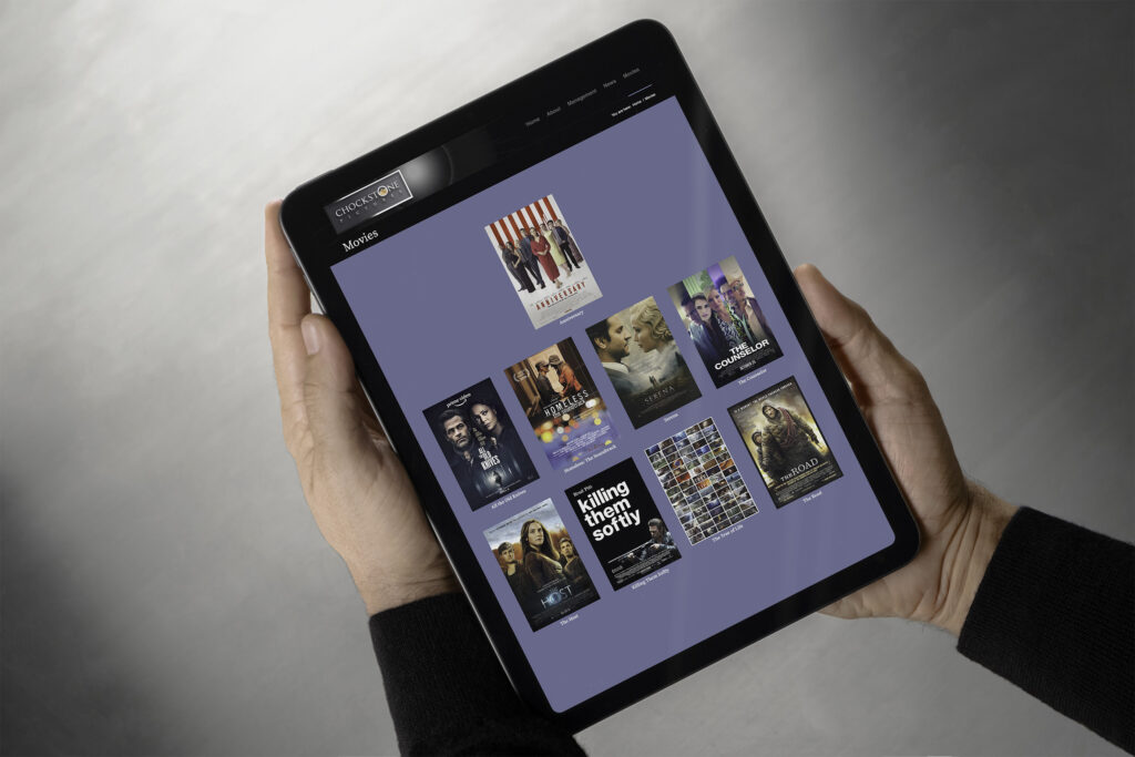 Hands hold a tablet flat over a gray backdrop showing the Chockstone Movies page, film posters from Serena and The Counselor to Killing Them Softly arranged as cards on slate blue.