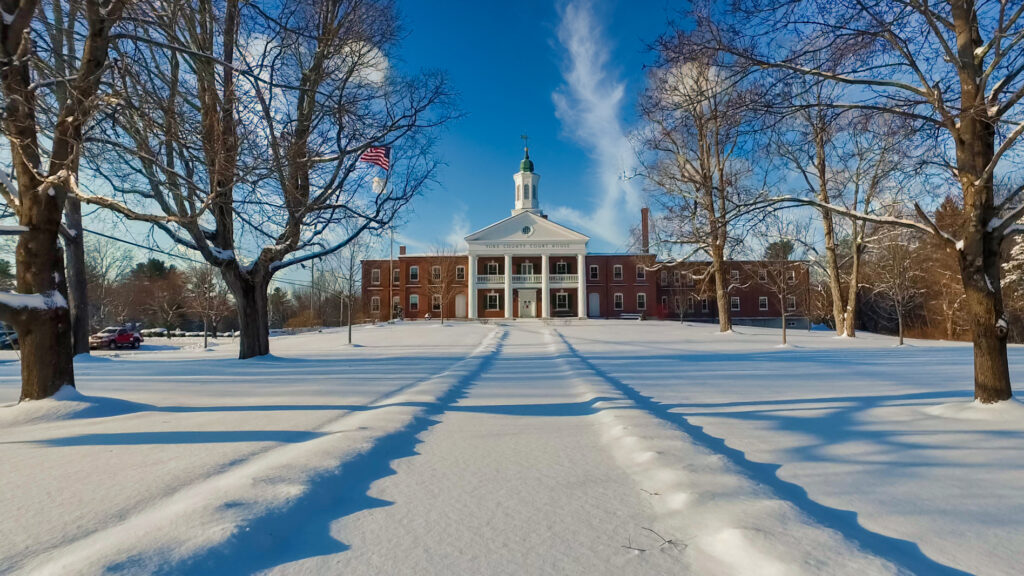 York County Courthouse in winter, the brick facade, white portico, and cupola rising behind an American flag across an unbroken field of snow.