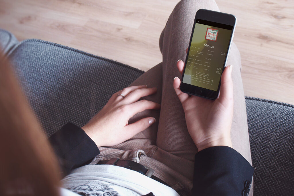 Woman on a gray couch holding a phone open to the Shows page, tour dates listed under the band mark.
