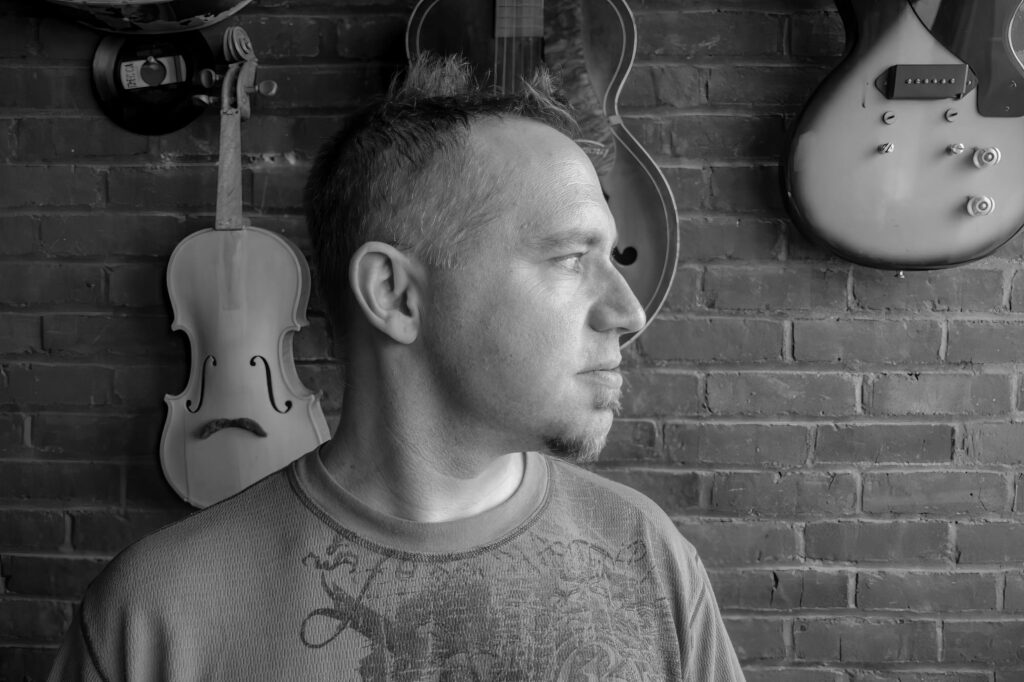 Profile portrait of a band member with a goatee against a brick wall hung with a violin and electric guitars.