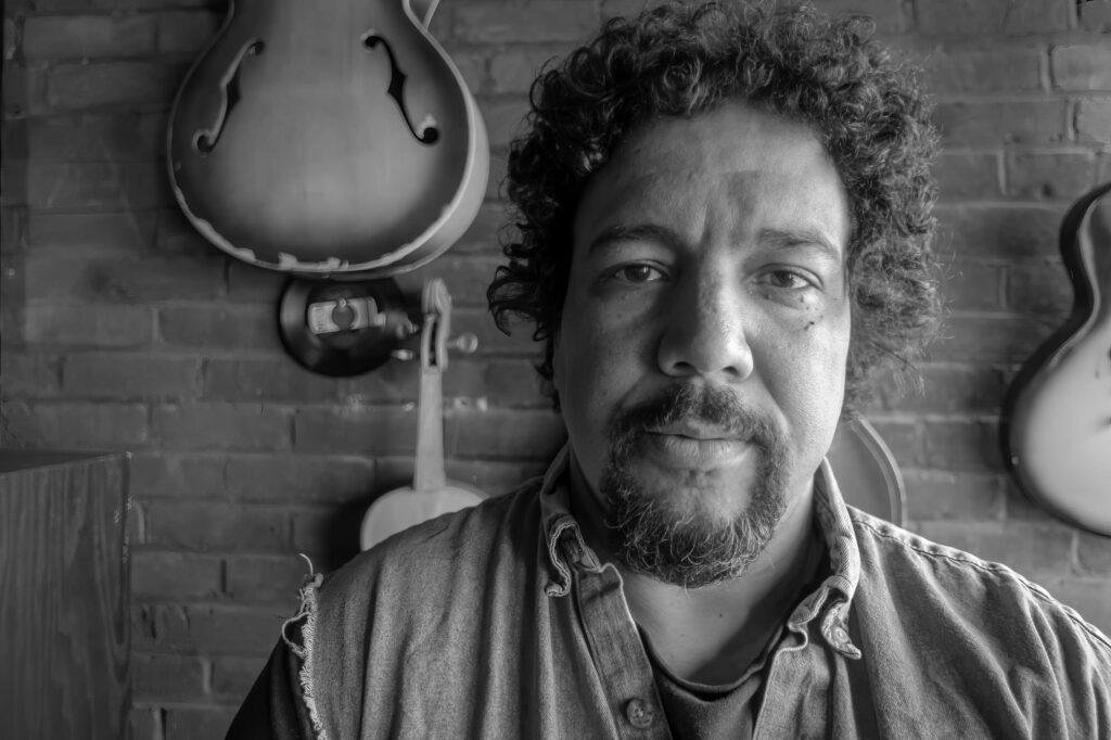 Close portrait of a curly-haired, bearded band member facing the camera, archtop guitar and violin hanging on the brick wall behind him.