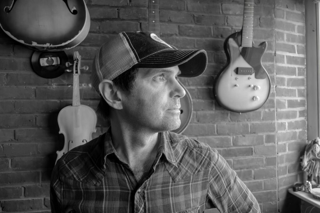 Band member in a mesh-back cap gazes past the camera, framed by a white violin and a single-cutaway electric guitar on brick.