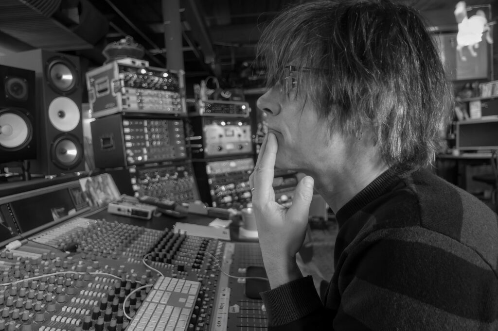 Engineer with long hair leans over a mixing console in thought, studio monitors and racks of outboard gear stacked behind him.
