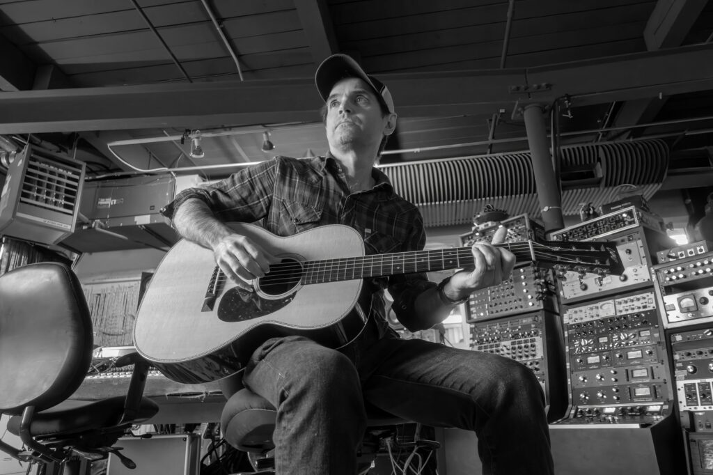 Guitarist in a plaid shirt and ball cap plays an acoustic guitar in the studio, surrounded by racks of vintage recording gear.