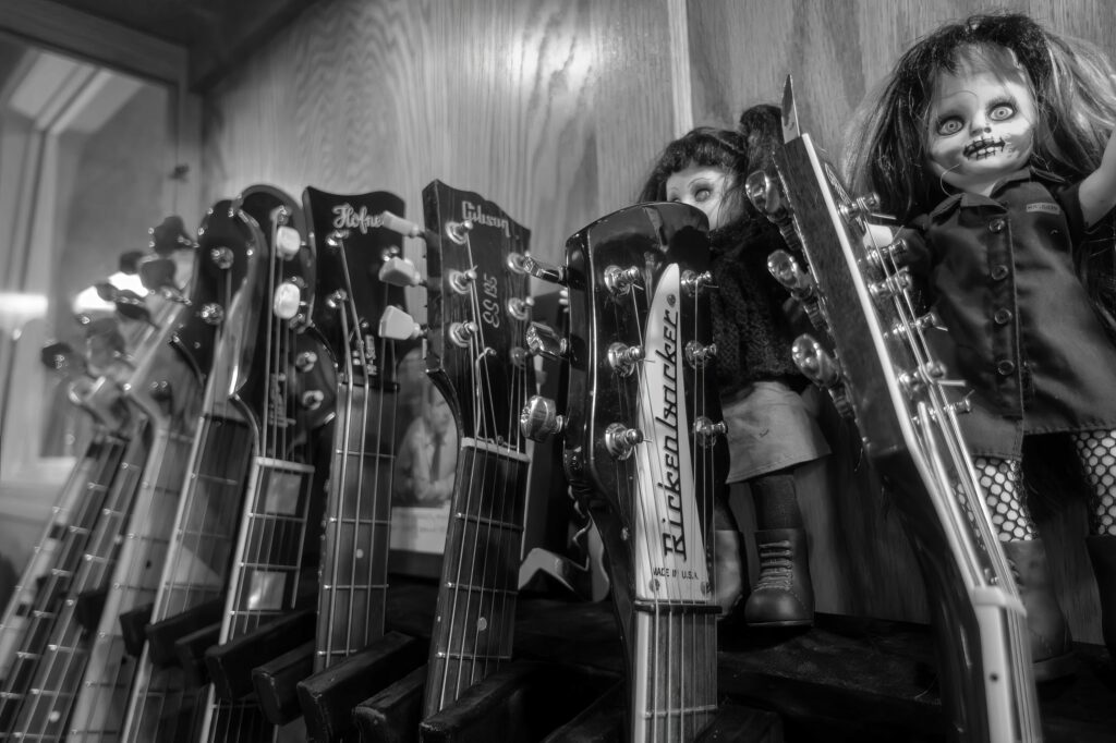 Lineup of Höfner, Gibson, and Rickenbacker headstocks in a display case, flanked by two gothic dolls peering between the guitars.