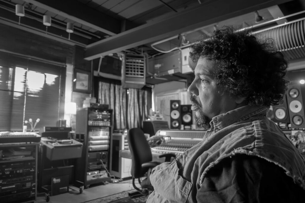 Curly-haired band member in profile inside the control room, window light falling across studio monitors and a mixing console.