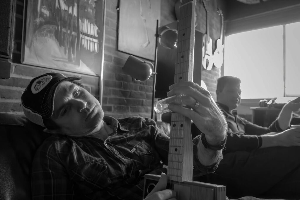 Musician in a trucker cap reclines on a couch cradling a cigar-box guitar neck while a bandmate talks near the window.