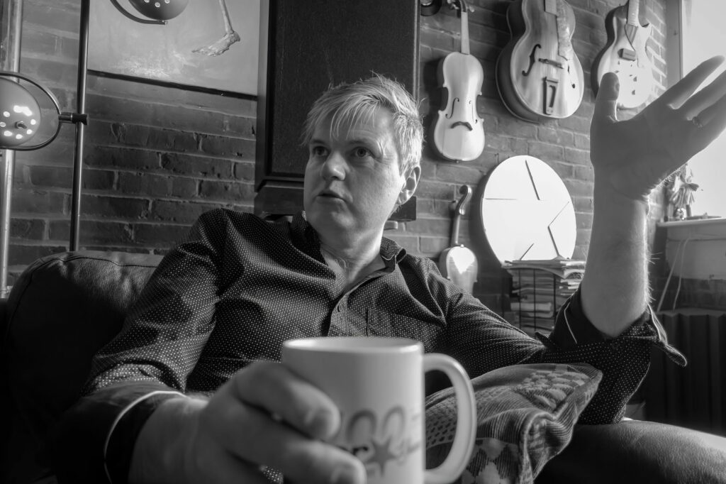 Band member gestures mid-conversation with a coffee mug in hand, white violin and archtop guitars hanging on the brick wall.