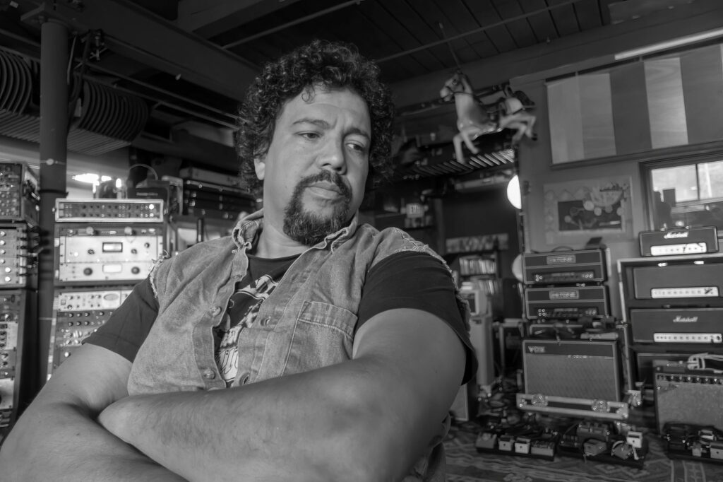 Band member with crossed arms and a goatee stands before stacked Marshall amps, a carousel horse perched above the studio gear.
