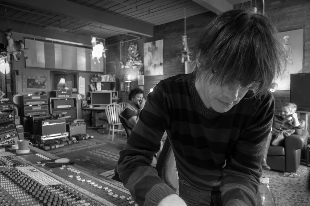 Producer in a striped sweater works over the mixing console while bandmates relax on chairs and a couch across the live room.