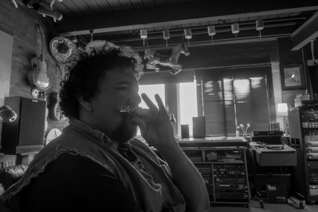 Backlit musician laughs while raising a harmonica by the studio window, instruments and folk-art figures lining the wall behind him.