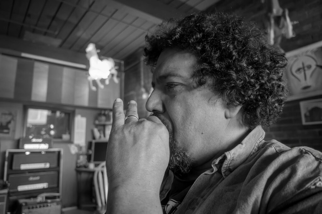 Close profile of a curly-haired musician playing harmonica, hands cupped to his mouth beneath a glowing horse-figure lamp.