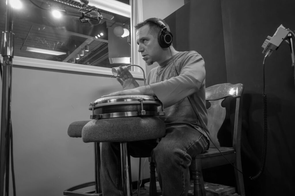 Percussionist in headphones plays a snare-mounted drum beside a studio window, microphone stands and a wooden chair framing the isolation booth.