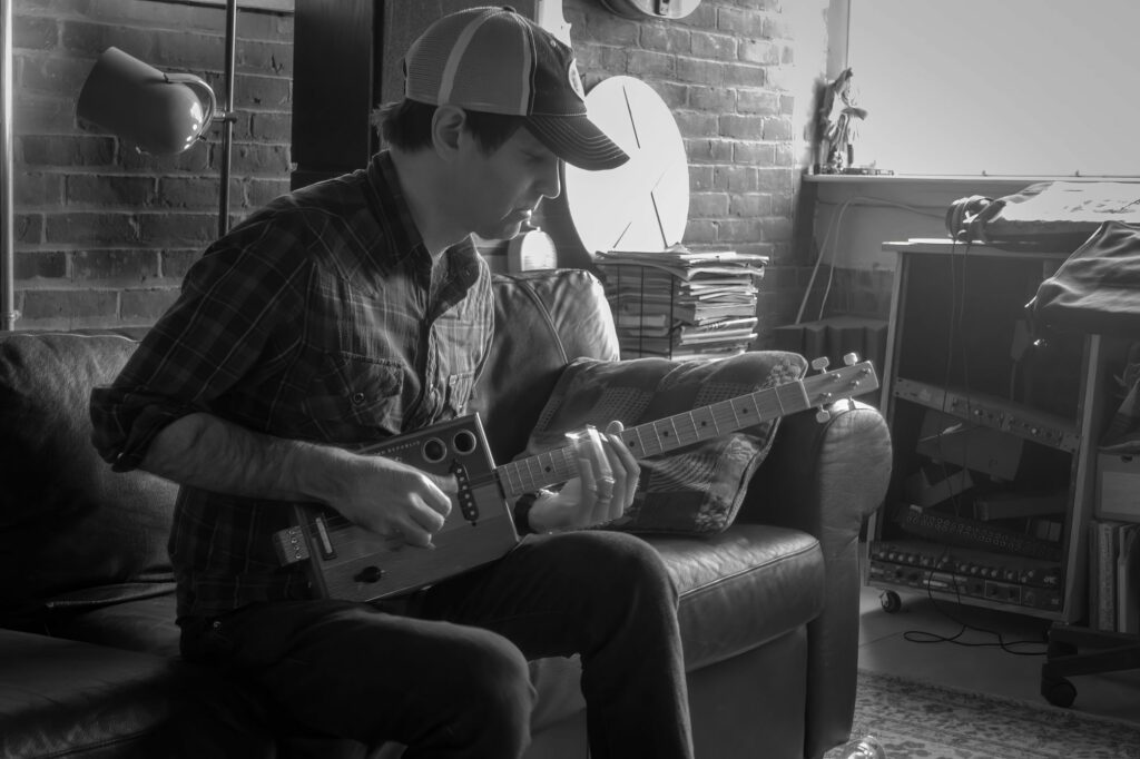 Musician in a trucker cap bends over a cigar-box guitar on a leather couch, brick walls and stacked records behind him.