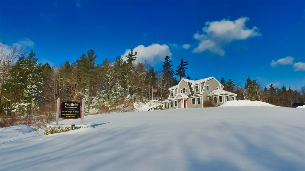 Fairfield & Associates’ gambrel-roofed Lyman office in deep snow, roadside sign and American flag standing before the gray shingled building with its red door.