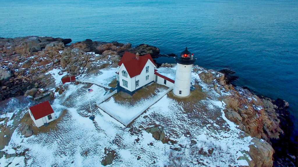 Aerial drone view of Nubble Light in winter daylight, red-roofed keeper’s house and lit lighthouse tower on snow-dusted rocks over teal water.