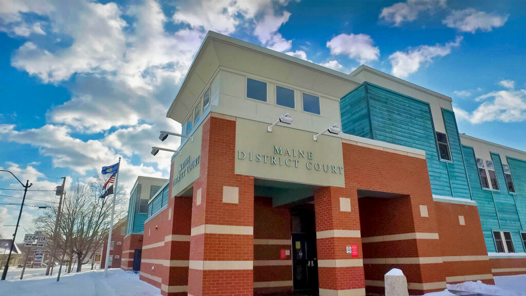 Maine District Court building in winter, its brick entrance tower and teal-clad wings rising under a bright blue sky with flags at the corner.