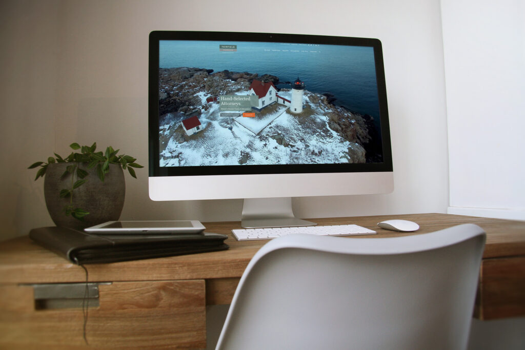 Fairfield & Associates homepage on an iMac in a warm wood office, the Nubble Light aerial hero introducing Hand-Selected Attorneys above the fold.