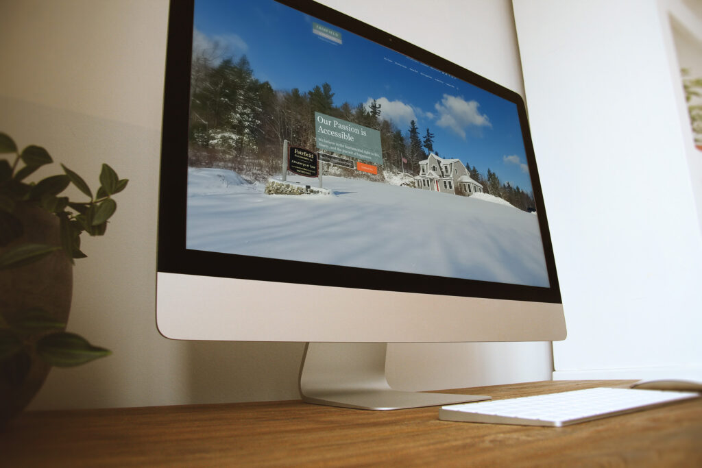 Fairfield & Associates website on an angled iMac, a snowy Maine hero scene carrying the Our Passion is Accessible message with an orange call to action.