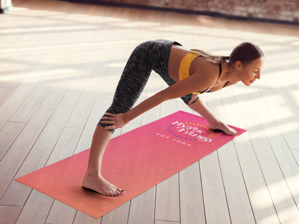 A student in a yellow top and gray leggings folds forward into a wide-legged stance, one hand on a coral Mystic Fitness yoga mat in a sunlit wood-floored studio.