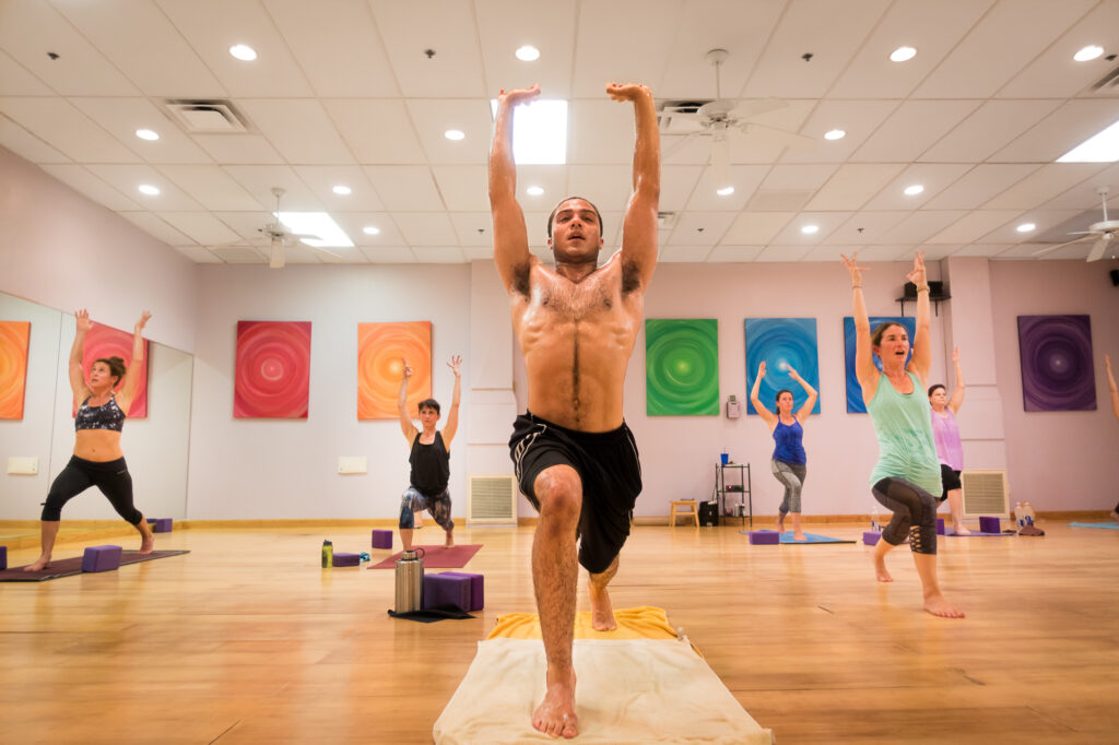 A student holds a one-legged standing balance with arms raised overhead in the sunlit Mystic Fitness studio, other practitioners behind.