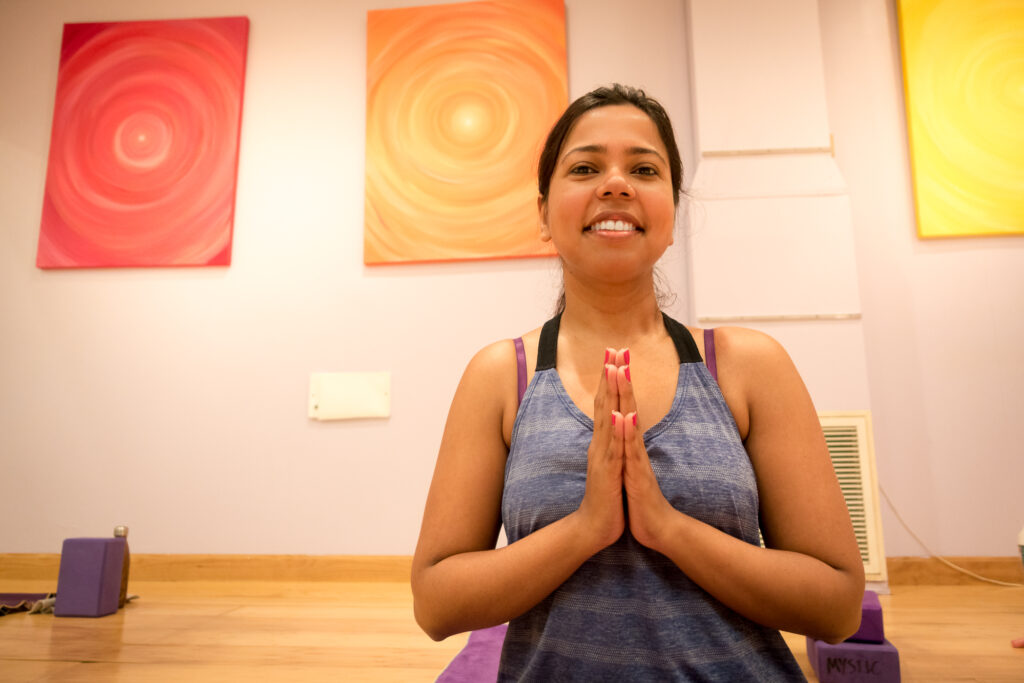 A student sits cross-legged with palms together at the chest, smiling, against the warm cream-and-orange studio wall.