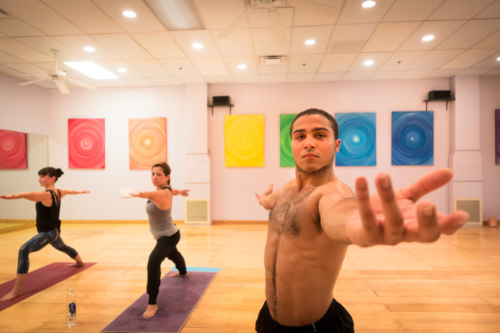 A student in warrior pose reaches an open hand toward the camera at the front of his mat, two classmates holding warrior behind him.