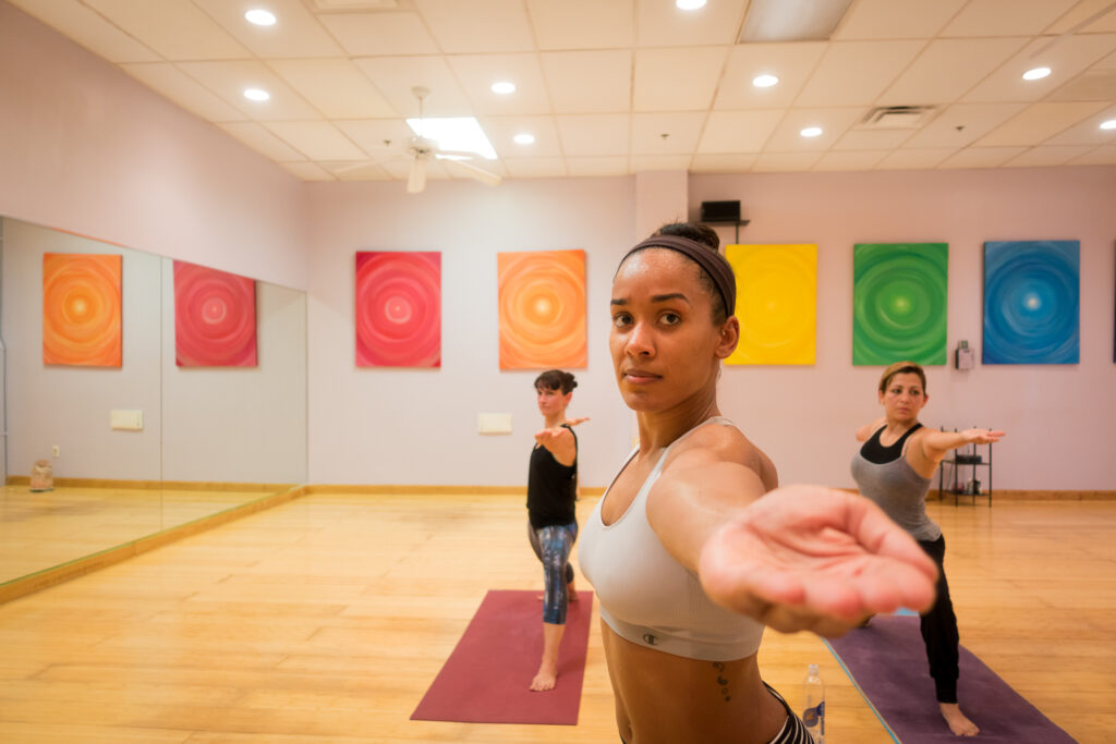 A student in a white sports top holds warrior pose and reaches an open hand toward the camera, two classmates in warrior behind her.