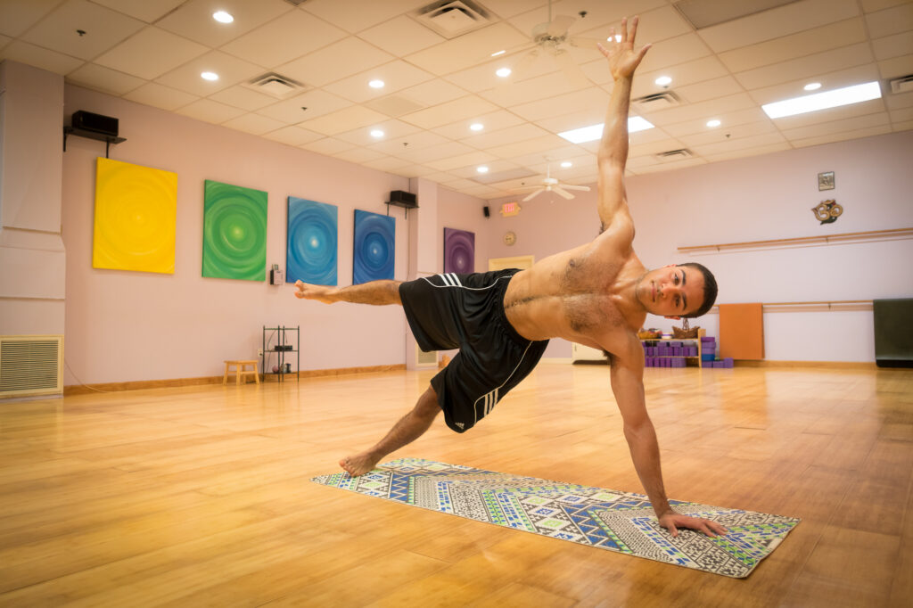 A student balances in a dynamic one-legged extended pose on the wood studio floor as warm light fills the mirrored room.