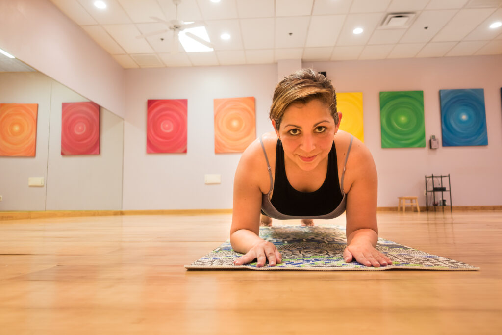 A student in a plank pose at the front of her mat, facing the camera in the warm studio.