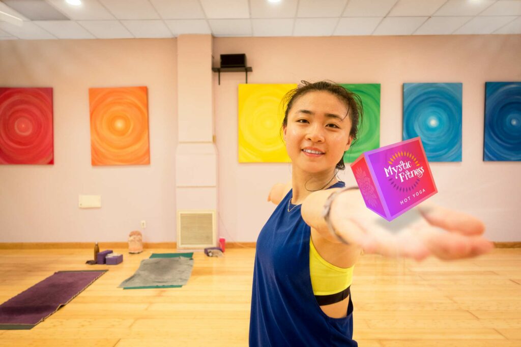A smiling student in a blue tank holds out the small Mystic Fitness cube on her open palm in the mural-lined studio.