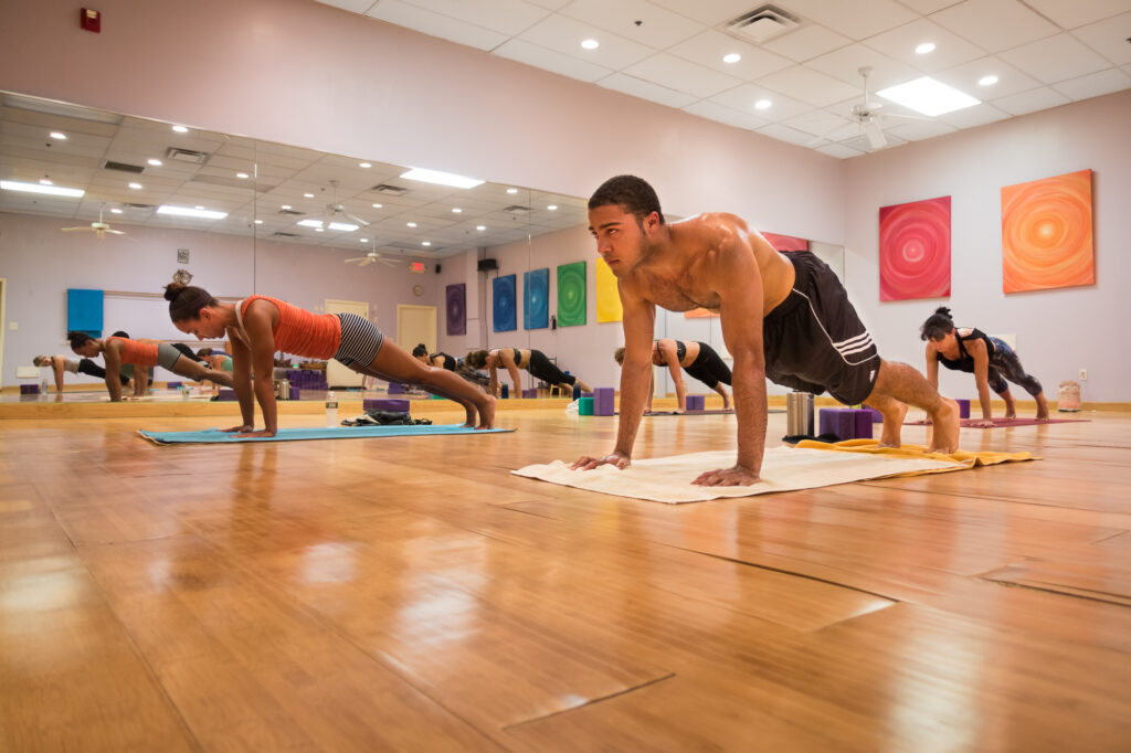 A row of students holds a plank pose across their mats in the bright, mirror-lined studio.