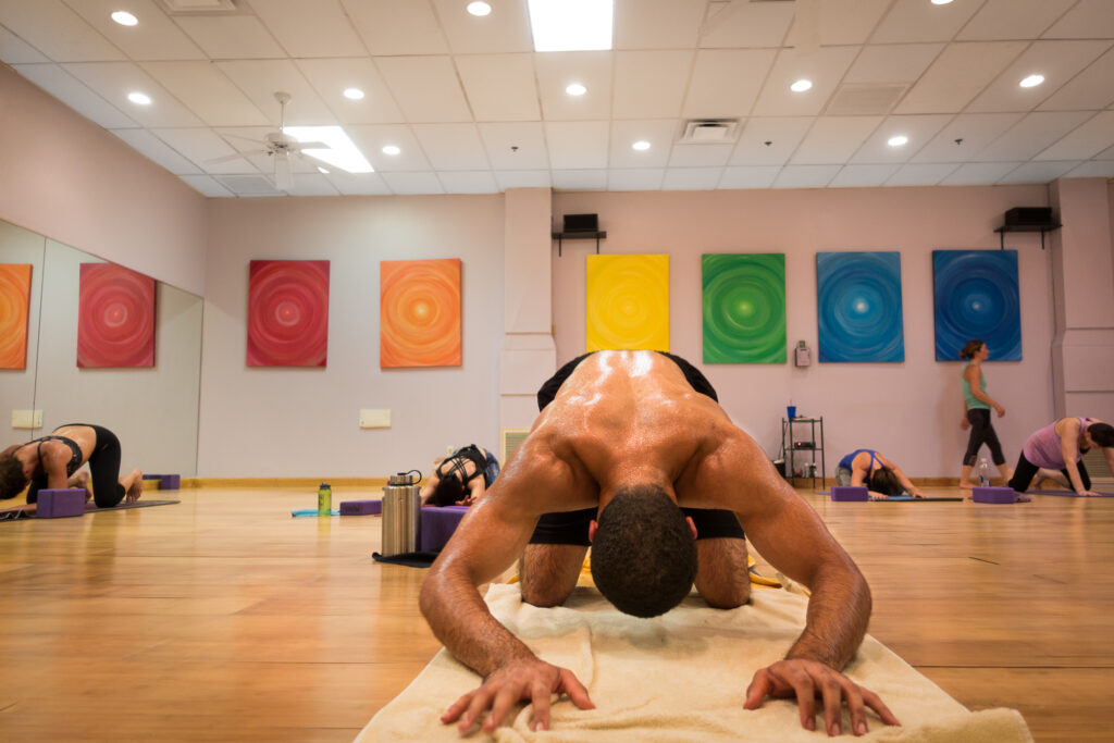 A student in a wide-legged forward fold, head near the floor and hands flat on his mat, with classmates in the same fold behind him.