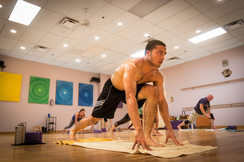 A student crouches in a low runner's lunge on his mat, hands to the floor, as classmates stretch behind him in the bright studio.