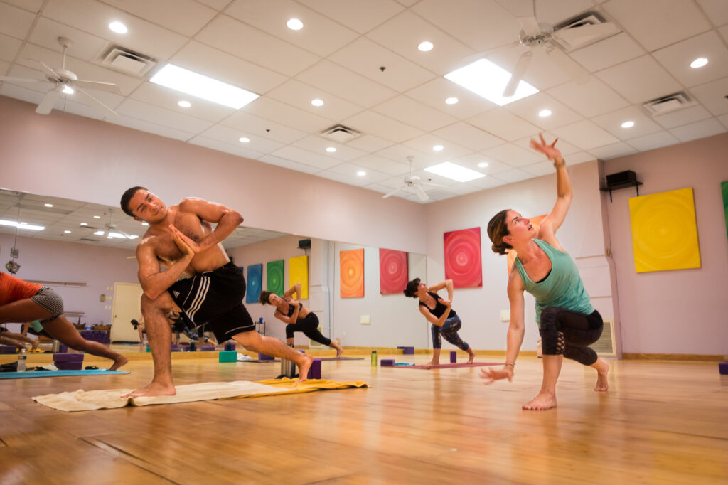 A student holds a low lunge in the foreground while classmates practice behind in the warm, mirror-lined studio.