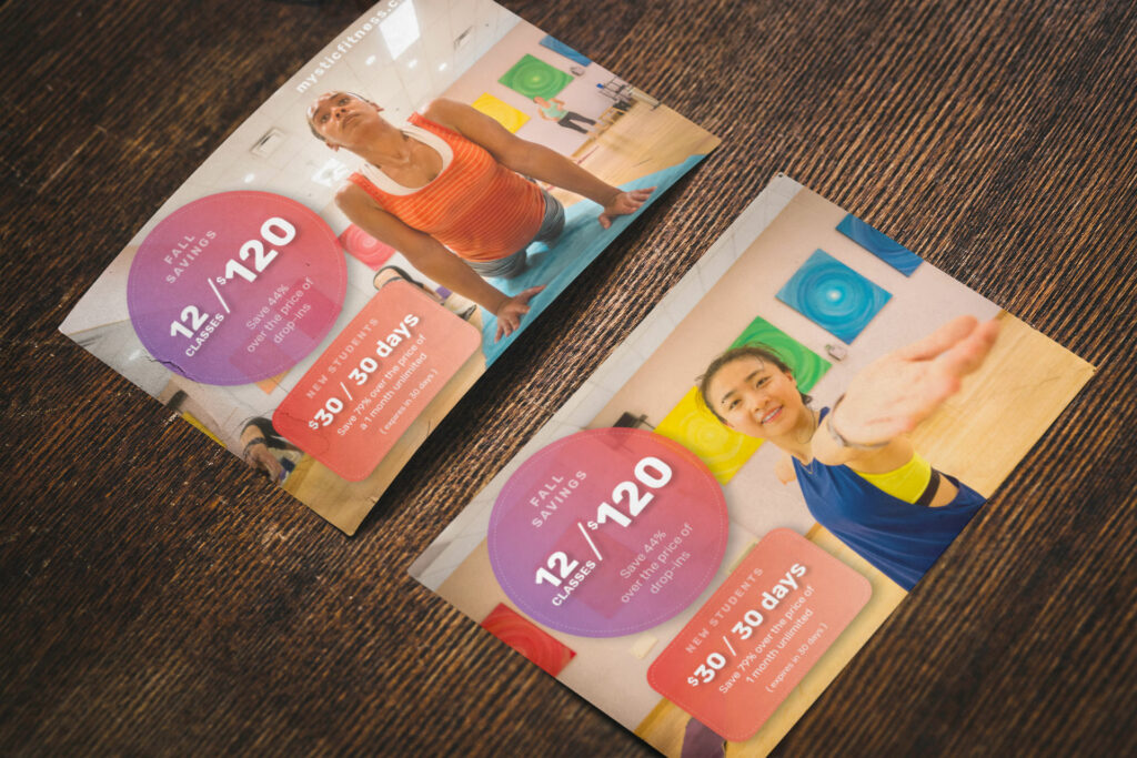 Two Fall Savings postcards on a dark wood table, one with a student in upward dog and one with a student holding the Mystic Fitness cube, both carrying the 12 classes for $120 offer.