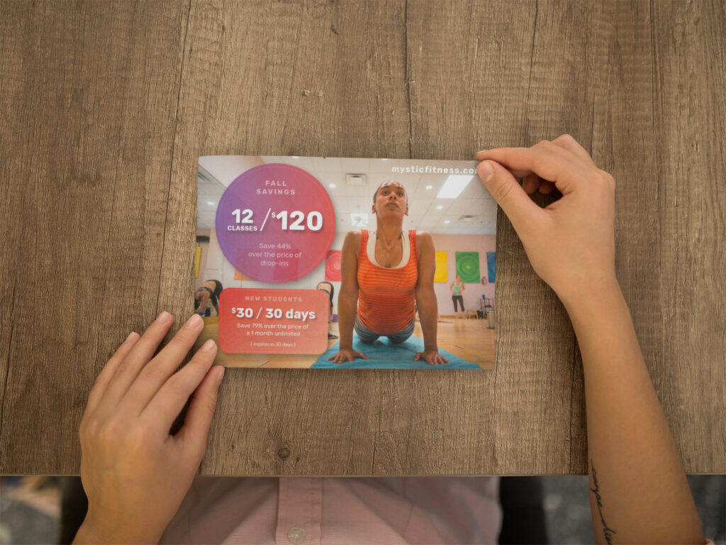 Hands place a Fall Savings postcard on a wooden table, a student in upward dog beside the 12 classes for $120 and $30 for 30 days offers.