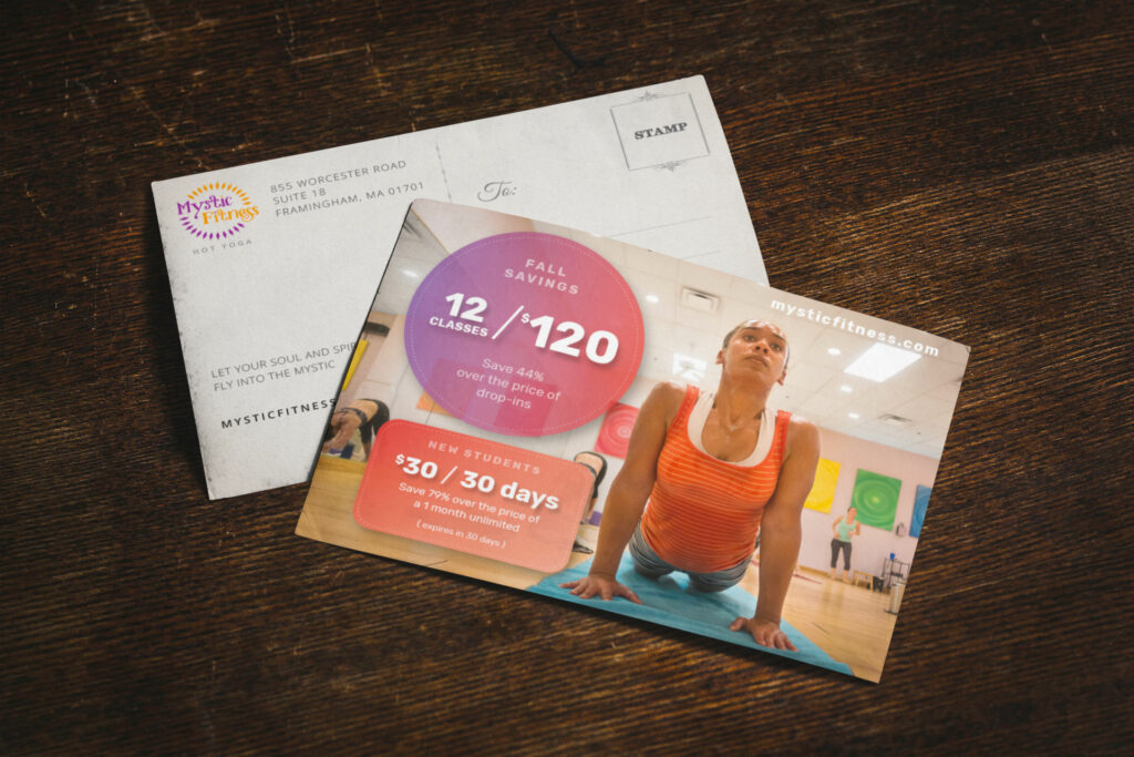 Front and address side of the Fall Savings postcard on dark wood, the back carrying the Mystic Fitness Hot Yoga logo, Framingham address, and stamp box.