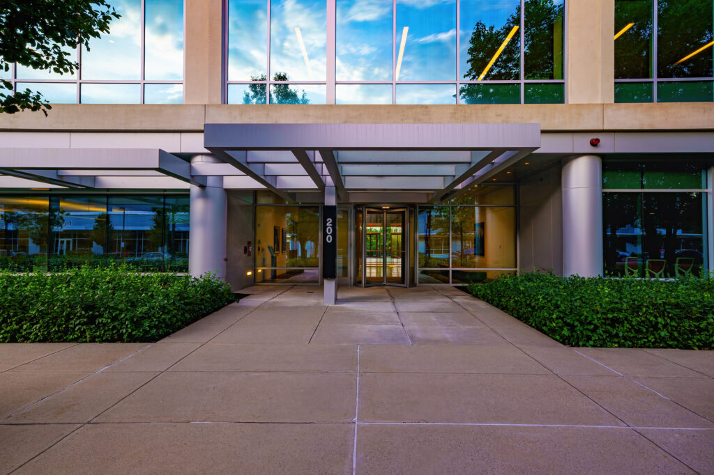 Concrete entry canopy and glass doors of the office building at dusk, evening sky reflected across the curtain wall.