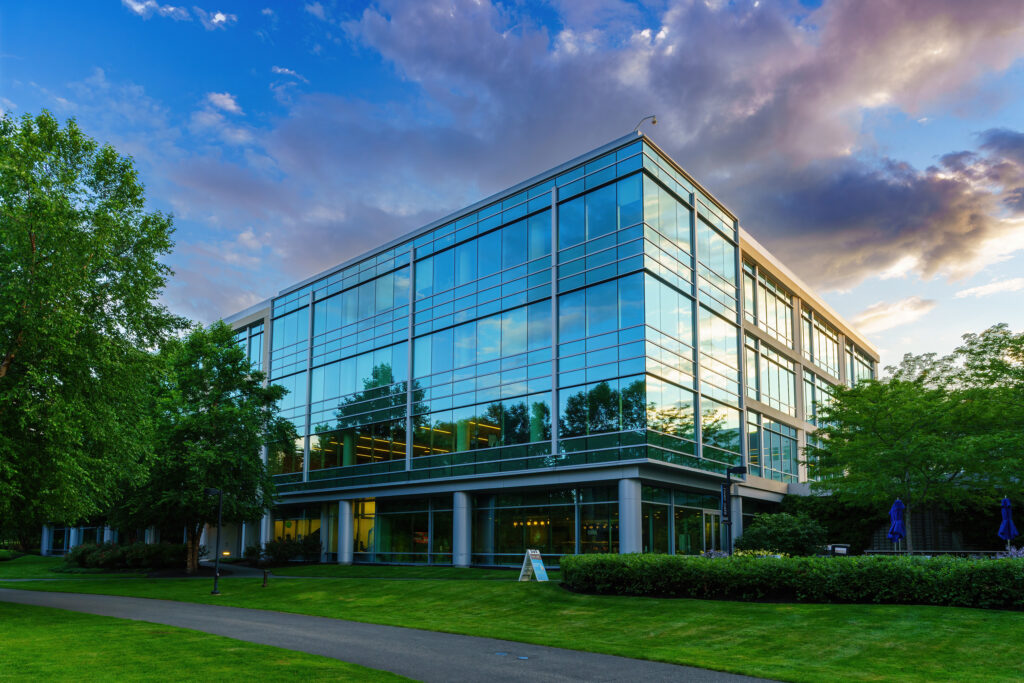 Corner view of the four-story glass office building at sunset, dramatic clouds reflected in the curtain wall above the lawn.