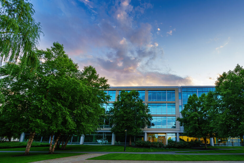 Front elevation of the glass office building behind summer trees, evening clouds drifting over the wide lawn.