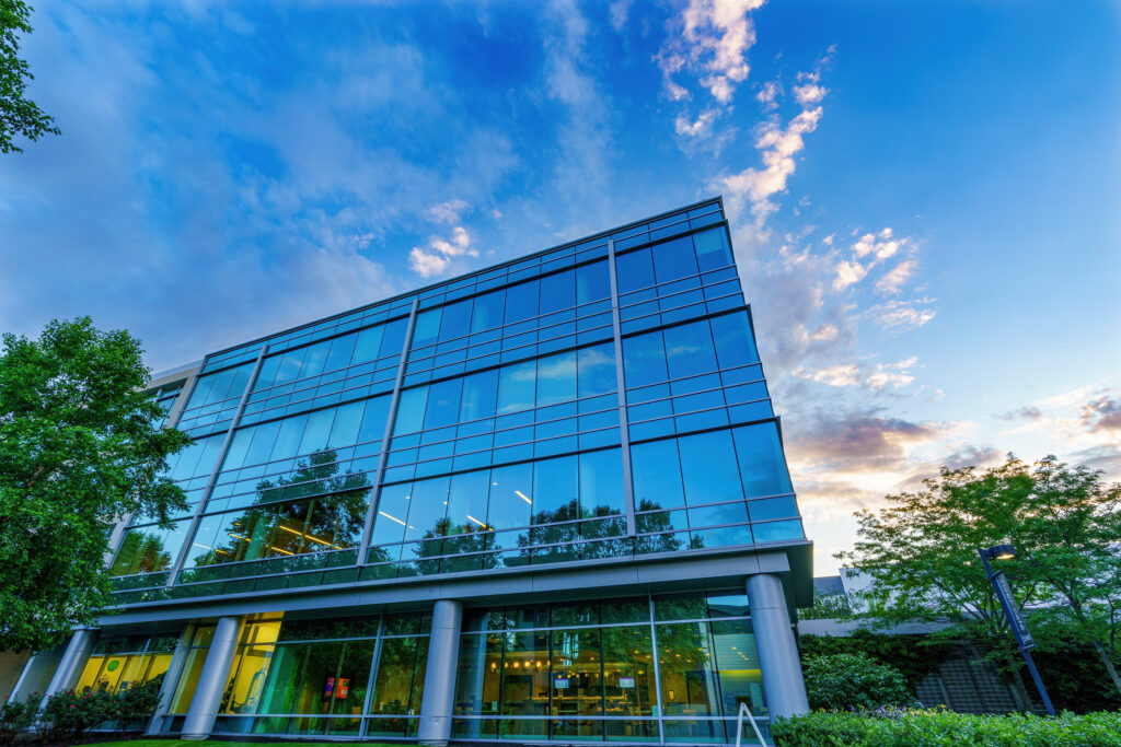 Low angle up the glass facade at dusk, warm office lights glowing along the ground floor beneath sunset clouds.